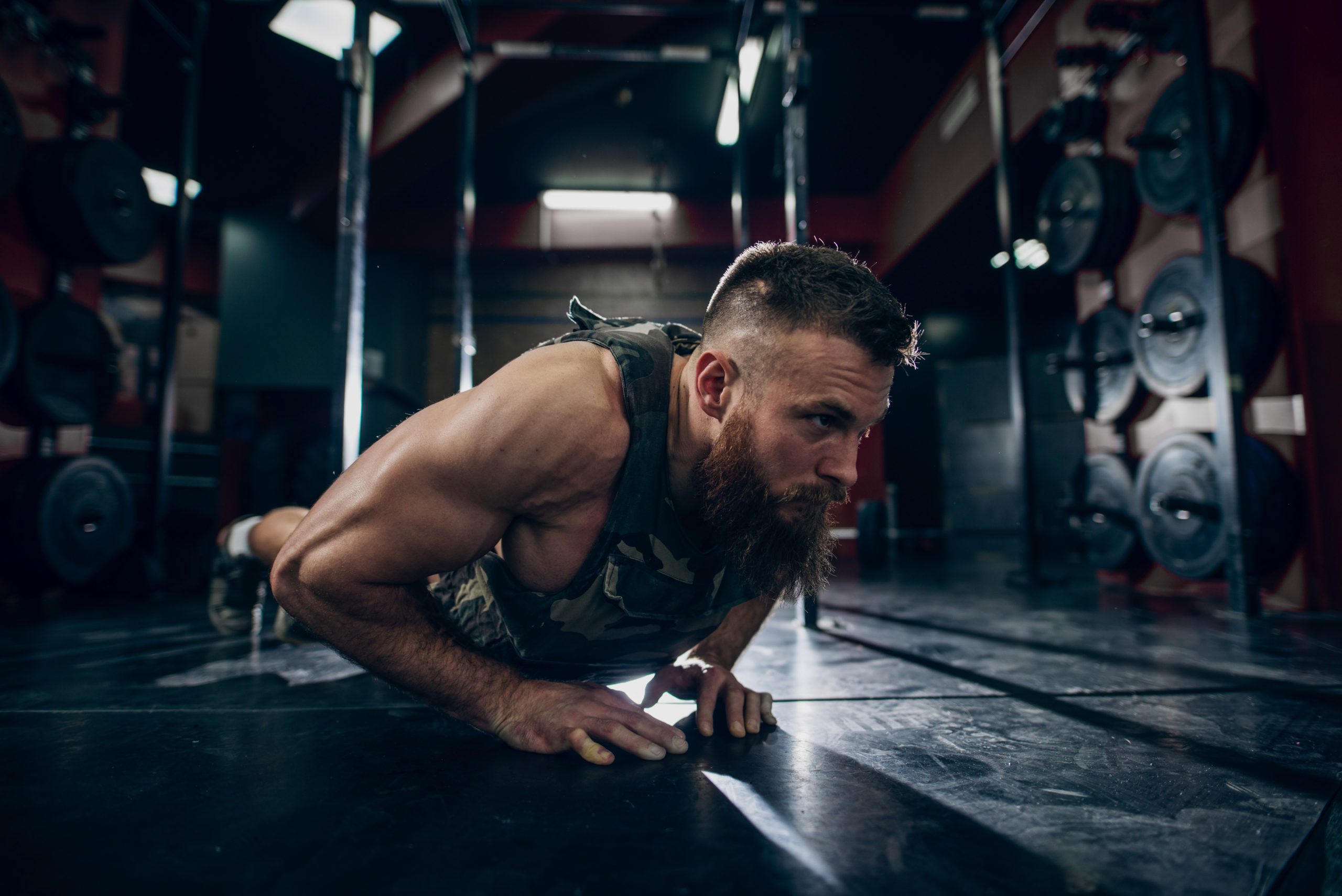 Muscular man doing push-ups in military style weighted vest in gym. Weight plates in background.