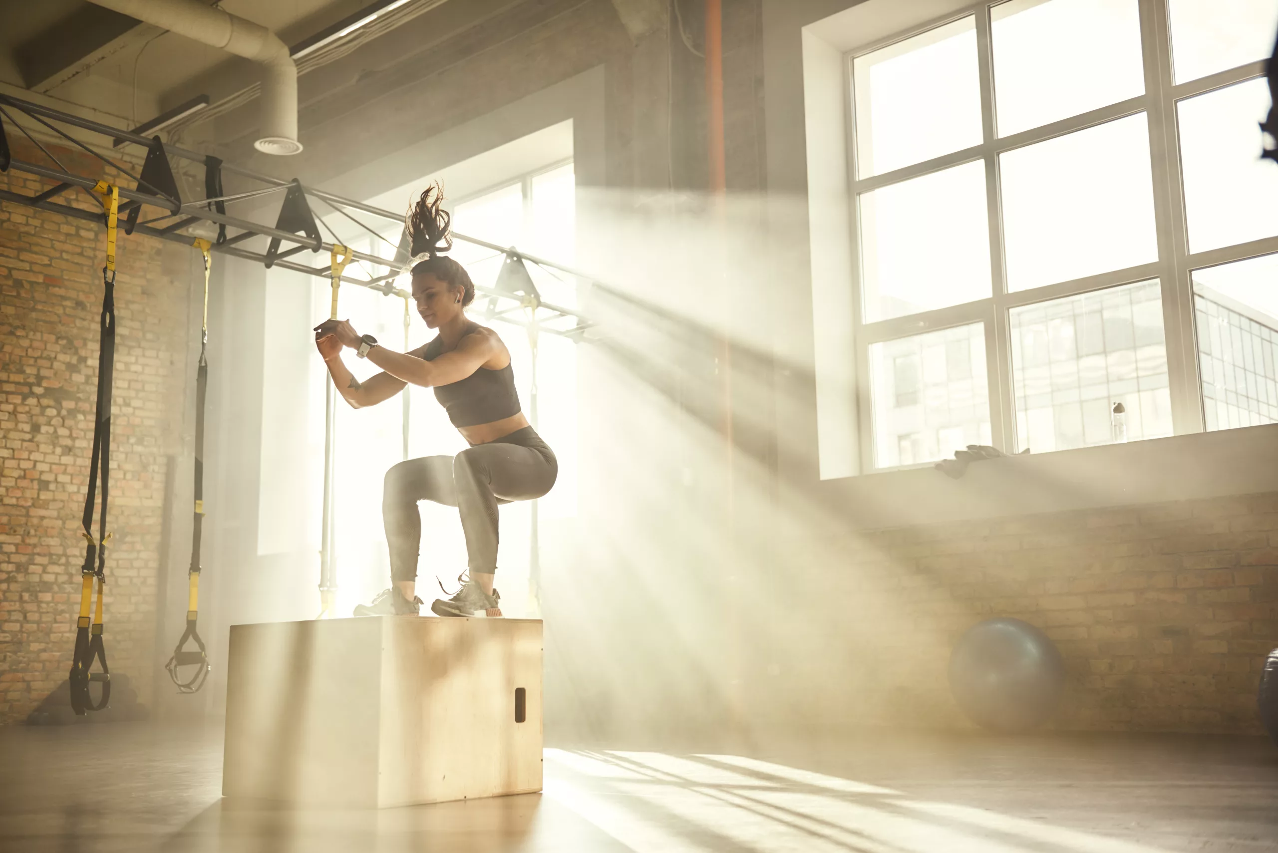 Side view of athletic woman in sportswear doing a box jump in gym with sun setting in window behind her