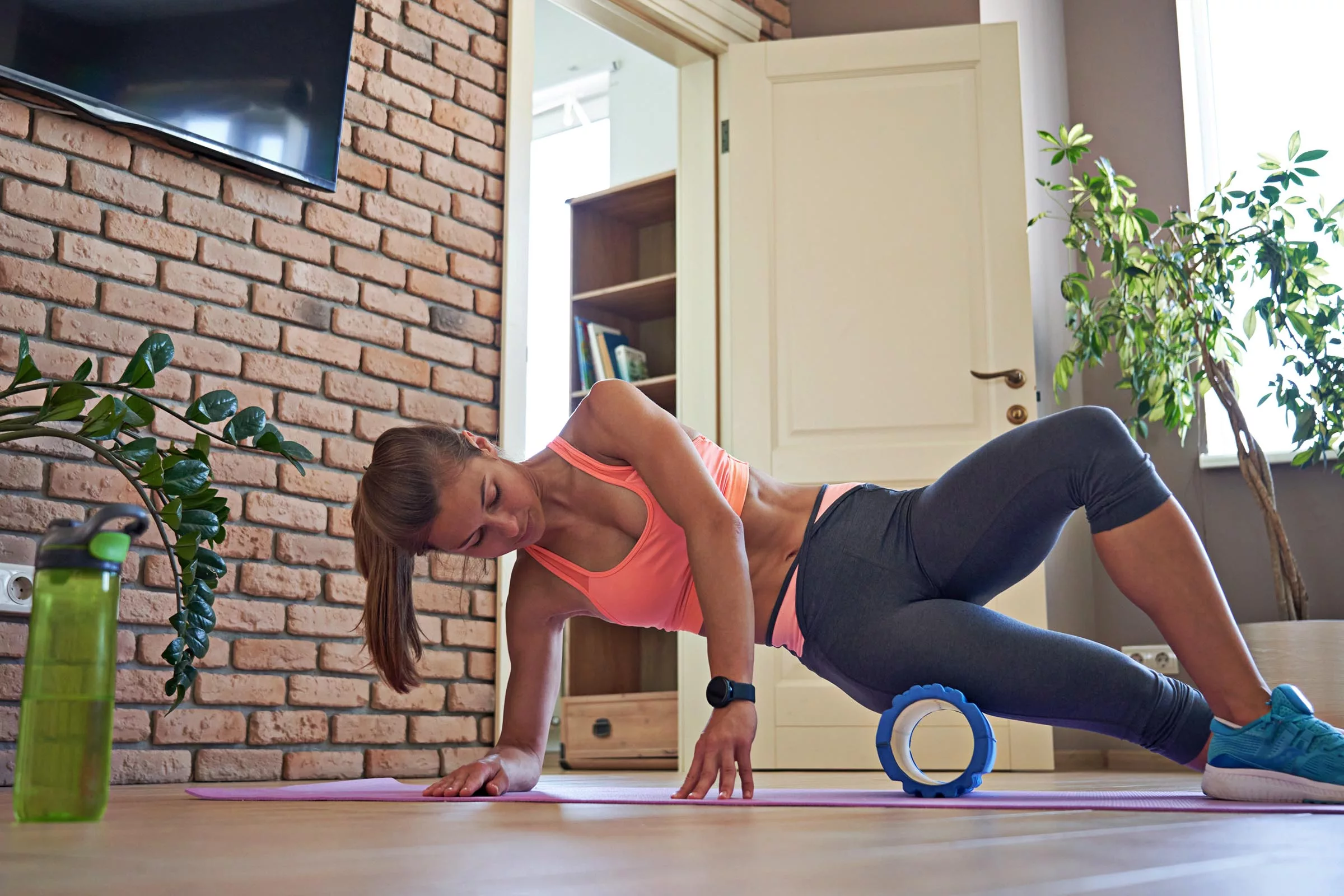 Woman athlete wearing sportswear using foam roller for muscle recovery on living room floor.