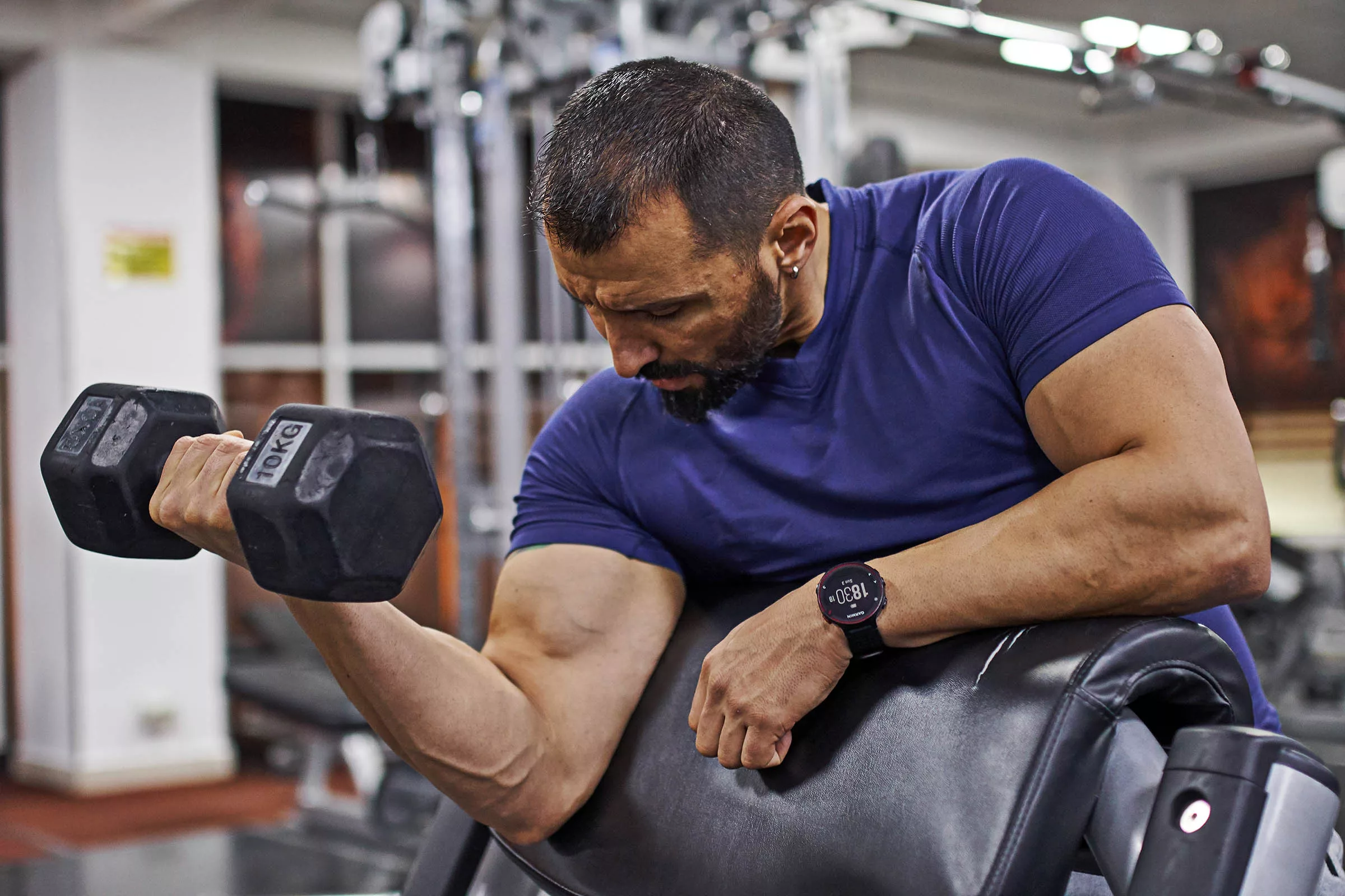 Man doing single-arm dumbbell preacher curl in gym for bigger biceps. 
