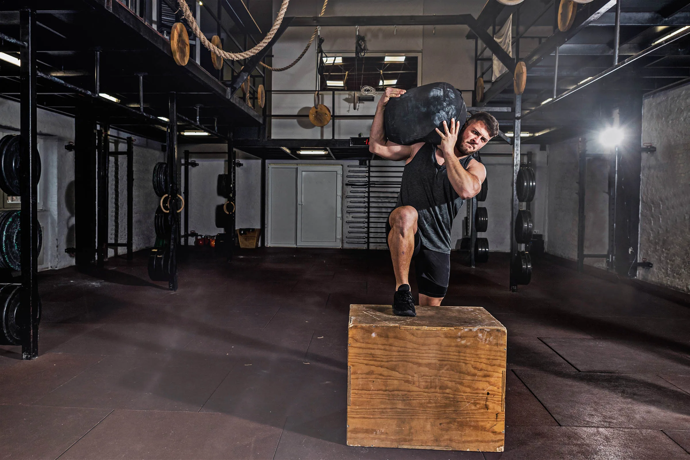 Man in functional fitness gym doing a sandbag workout on top of a box for full-body strength.