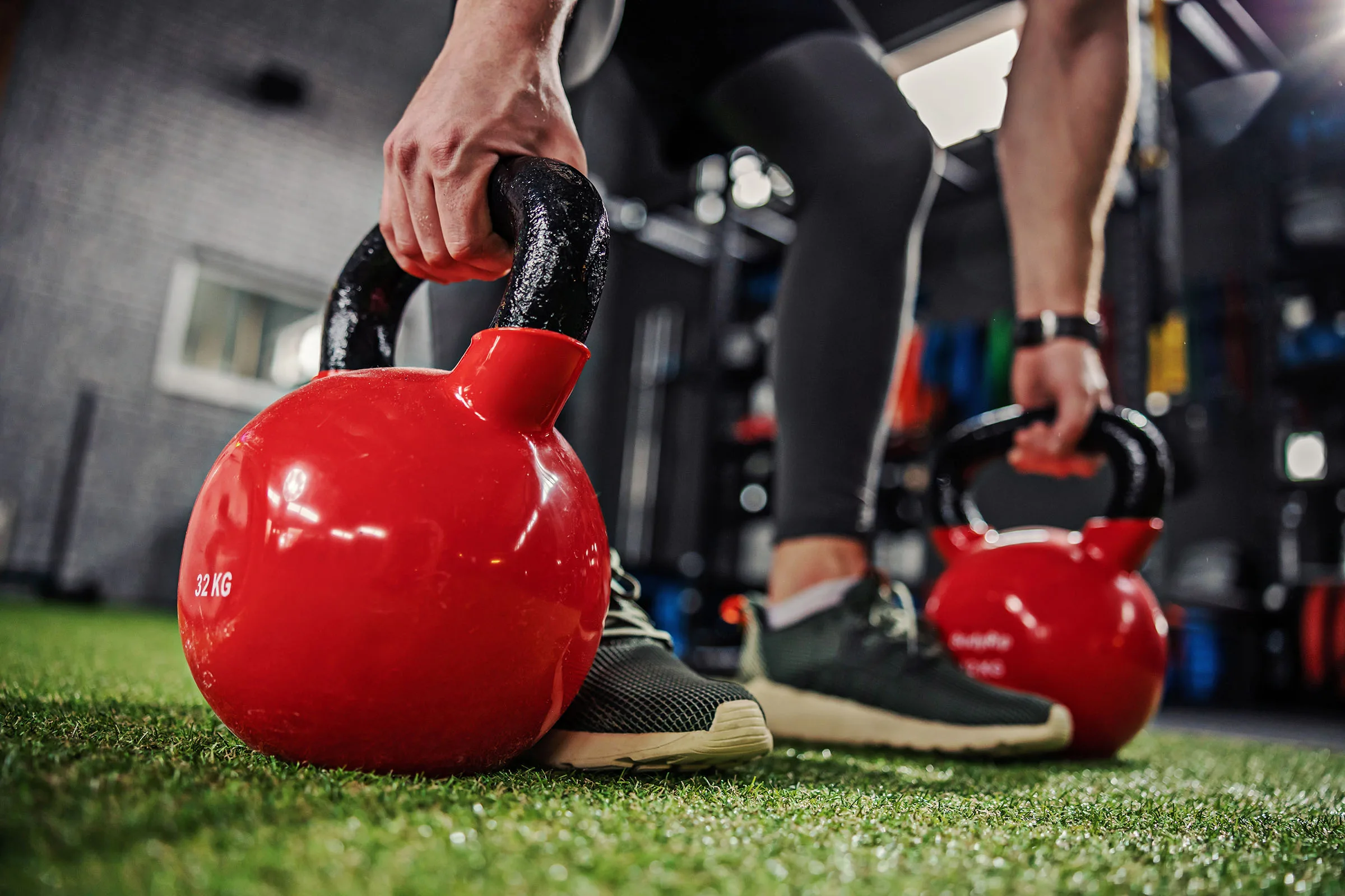 Kettle bells in focus. Two masculine strong arms take the sports equipment for exercising grip strength in both arms and prepare to lift weights. Two red kettle bells on green artificial grass indoor modern gym