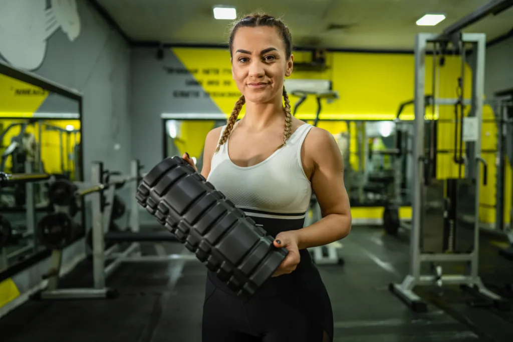 Female athlete standing in gym looking straight at camera while holding a foam roller