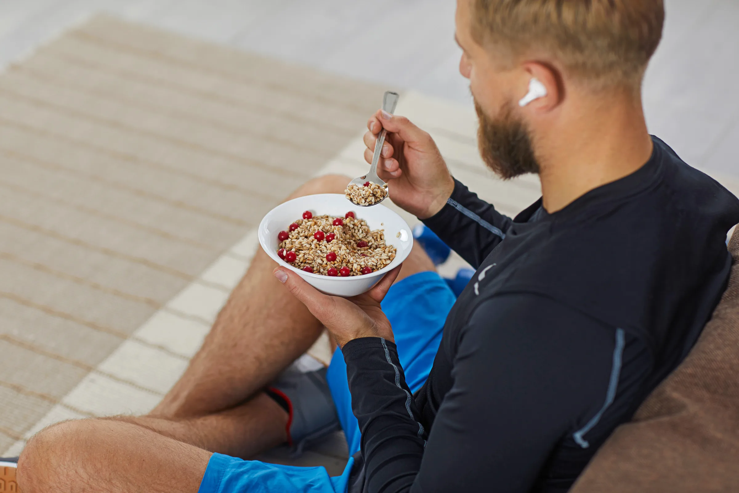 Young male athlete is having a bowl of healthy granola after his fitness workout kettlebells in green grass - fitness concept outdoors